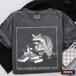 May include: Charcoal grey sweatshirt with a graphic of a raccoon in sunglasses toasting a marshmallow over a fire on books. The design is framed in white, with "DR. WILSON - 2026" printed below.