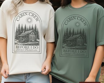 May include: Two t-shirts, one ivory and one moss green, featuring a cabin graphic with text. The graphic includes a cabin, trees, sun, and the words "CABIN CREW" and "BEFORE I DO Emily's Bachelorette".