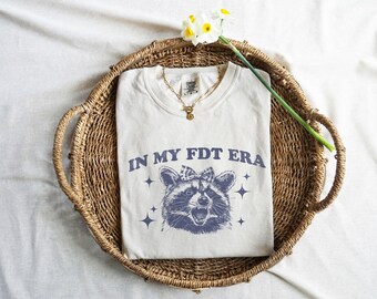 May include: White t-shirt with a blue raccoon graphic and the text "IN MY FDT ERA".