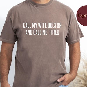 Puede incluir: Una camiseta marrón con el texto "Call my wife doctor and call me tired" impreso en blanco.