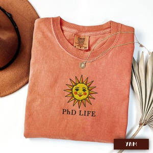May include: Coral-coloured t-shirt featuring a smiling sun graphic and the text "PhD LIFE". A gold necklace with a pendant rests on the shirt. A brown hat and dried palm leaf are in the background.