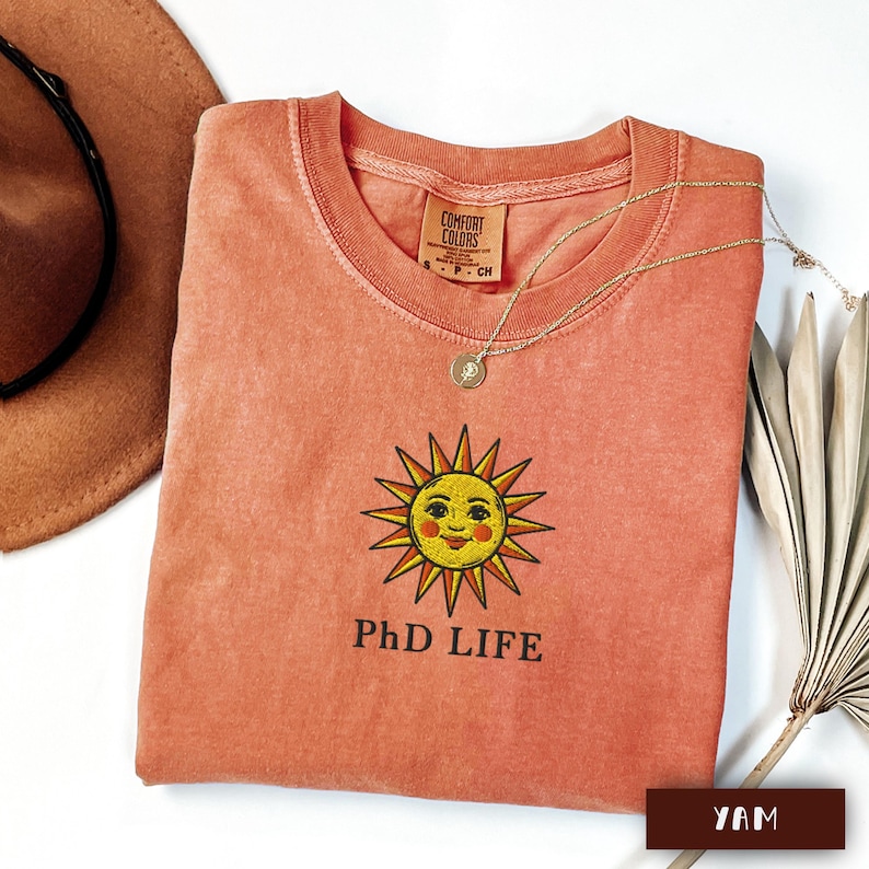 May include: Coral-coloured t-shirt featuring a smiling sun graphic and the text "PhD LIFE". A gold necklace with a pendant rests on the shirt. A brown hat and dried palm leaf are in the background.