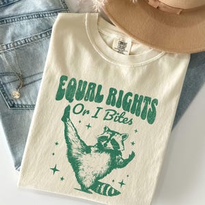 Puede incluir: Camiseta color crema con el texto "EQUAL RIGHTS Or I Bites" en verde, con un gráfico de mapache. La camiseta está colocada sobre unos vaqueros azules y un sombrero beige.