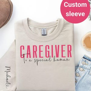 May include: A beige sweatshirt with the text "Caregiver to a special human" in pink and black lettering. The text "Michael" is written on the sleeve in black.