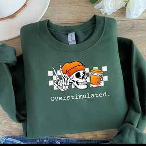 May include: Dark green sweatshirt with a graphic of a skull wearing an orange beanie, giving a peace sign with a skeleton hand, and holding a coffee cup. The text "Overstimulated" is below the graphic.