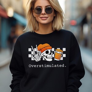 May include: Black sweatshirt with a graphic featuring a skull wearing an orange beanie, a peace sign, and a beer mug. The text "Overstimulated" is printed below the graphic.