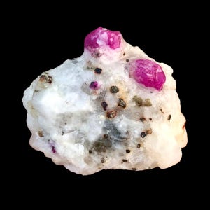 May include: A natural ruby specimen featuring a white base rock. Two bright pink rubies are prominent, with smaller ruby inclusions and dark mineral deposits. The background is black.