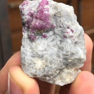 May include: A natural gemstone with a light grey matrix, hosting a cluster of bright pink crystals. The stone's rough, irregular form highlights its raw, unrefined beauty, typical of mineral specimens.