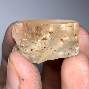 May include: A faceted, translucent crystal with a light amber hue. The crystal has a rectangular shape with multiple facets and small, reddish-brown inclusions. The edges are sharp, and the overall appearance is clear and well-defined.