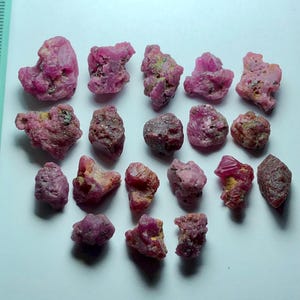 May include: A collection of rough, uncut ruby gemstones. The rubies vary in size and shape, displaying a range of pink and red hues. Some stones have a slightly rough, textured surface, while others appear more crystalline. The gemstones are scattered on a white surface.