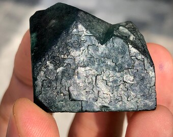 Könnte beinhalten: Ein dunkelblau-grünes Mineralexemplar mit rauer, strukturierter Oberfläche. Die Oberfläche weist ein mosaikartiges Muster mit helleren grauen Flecken auf. Die Kanten sind kantig und die Form ist grob rechteckig. Das Mineral wird in einer Hand gehalten.