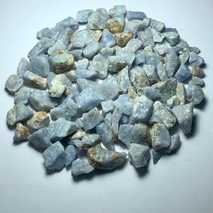 May include: A collection of small, irregularly shaped blue-gray stones with some tan and brown patches. The stones are scattered across a white surface, creating a natural, earthy aesthetic. These raw gemstones are suitable for jewelry making or decorative purposes.