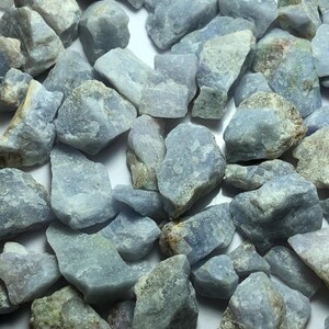 May include: A collection of rough, light blue gemstones. The stones vary in size and shape, with a mix of smooth and textured surfaces. Some stones have hints of gray and brown.