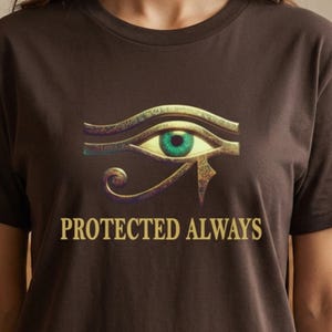 May include: A dark brown short-sleeve t-shirt featuring a graphic of the Eye of Horus in gold and green tones centered on the chest. Below the eye, the text "PROTECTED ALWAYS" is printed in a gold, serif font. The shirt has a crew neckline.