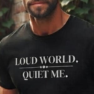 Op de afbeelding: Een zwart T-shirt met korte mouwen en de witte tekst LOUD WORLD. QUIET ME. op de borst gedrukt. Het shirt heeft een ronde hals en een standaard pasvorm, met een duidelijk grafisch ontwerp voorzien van een horizontale lijn en een ruitvormig accent.
