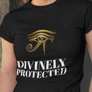 May include: A black short-sleeved t-shirt featuring a gold-colored Eye of Horus graphic centered on the chest. Below the graphic, the text DIVINELY PROTECTED is printed in white capital letters. The shirt has a crew neckline and a fitted silhouette.