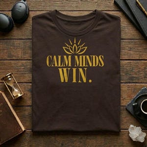May include: A dark brown t-shirt featuring a gold graphic of a lotus flower icon above the text "CALM MINDS WIN." The shirt has a crew neckline and short sleeves, presented as a flat lay garment with a centered, bold, sans-serif typographic design.