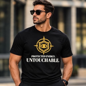 Op de afbeelding: Een zwart t-shirt met korte mouwen en een gouden afbeelding van een oog in een schild- en kompasontwerp. Onder de afbeelding staat de tekst PROTECTED ENERGY. UNTOUCHABLE. in witte hoofdletters. Het shirt heeft een ronde hals.