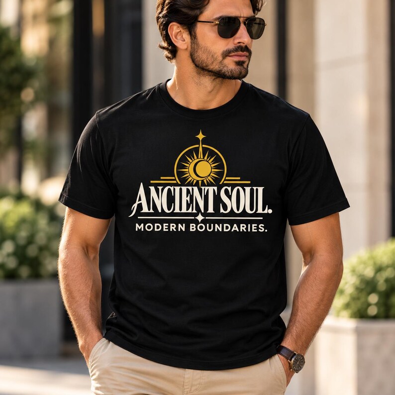 Op de afbeelding: Een zwart T-shirt met korte mouwen voorzien van een gouden afbeelding van een zon- en maansikkelpictogram boven de tekst ANCIENT SOUL. en de tekst MODERN BOUNDARIES. in het wit eronder gedrukt, gecentreerd op de borst van het kledingstuk.
