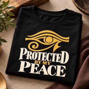 May include: A black t-shirt featuring a yellow graphic of the Eye of Horus above the white and yellow text that reads PROTECTED BY MY PEACE. The shirt has a crew neckline and is folded flat, displaying the central graphic design clearly.