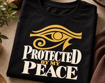 Può includere: Una t-shirt nera con una grafica gialla dell'Occhio di Horus sopra il testo bianco e giallo che recita PROTECTED BY MY PEACE. La maglietta ha uno scollo rotondo ed è piegata in piano, mostrando chiaramente il design grafico centrale.