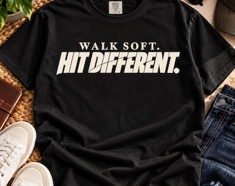 Può includere: Una t-shirt nera a maniche corte con il testo bianco "WALK SOFT." sopra il testo bianco più grande "HIT DIFFERENT." con una barra diagonale attraverso la parola "DIFFERENT.". La maglietta ha un girocollo e un'etichetta Comfort Colors.