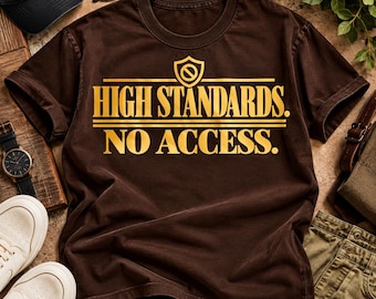 Può includere: Una t-shirt marrone a maniche corte con una stampa grafica dorata sul petto. Il testo recita "HIGH STANDARDS. NO ACCESS." con un'icona a scudo contenente un simbolo di divieto centrato sopra le parole, tutto in un carattere grassetto.