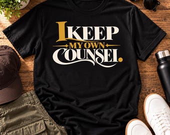 Può includere: Una t-shirt nera a maniche corte con il testo I KEEP MY OWN COUNSEL stampato in un carattere stilizzato color oro e crema sul petto. Il design include accenti a forma di stella decorativa e un piccolo punto dorato dopo l'ultima parola.