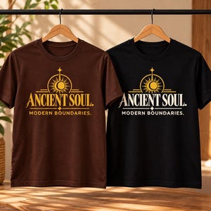 Op de afbeelding: Twee t-shirts met korte mouwen, &eacute;&eacute;n bruin en &eacute;&eacute;n zwart, elk voorzien van een gouden grafische print van een zon- en maansymbool boven de tekst ANCIENT SOUL en MODERN BOUNDARIES. De shirts hangen aan houten kledinghangers aan een zwart rek.