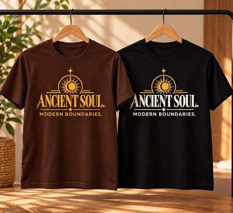 Op de afbeelding: Twee t-shirts met korte mouwen, &eacute;&eacute;n bruin en &eacute;&eacute;n zwart, elk voorzien van een gouden grafische print van een zon- en maansymbool boven de tekst ANCIENT SOUL en MODERN BOUNDARIES. De shirts hangen aan houten kledinghangers aan een zwart rek.