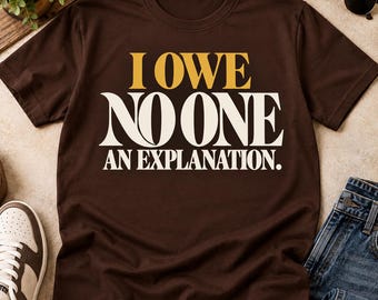 Può includere: Una t-shirt a maniche corte marrone scuro con il testo I OWE NO ONE AN EXPLANATION stampato sul davanti. La parola I OWE è in oro, mentre le parole NO ONE AN EXPLANATION sono in bianco, tutte in un carattere tipografico con grazie.