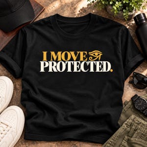 May include: A black short-sleeved t-shirt featuring the text "I MOVE" in gold lettering followed by an Eye of Horus symbol, with the word "PROTECTED." printed in large white capital letters underneath on the front of the garment.