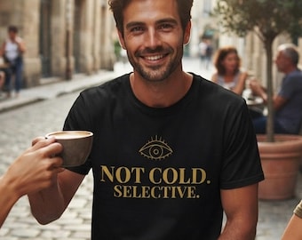 Può includere: Una t-shirt nera a maniche corte con una grafica dorata di un occhio sopra il testo "NOT COLD. SELECTIVE." in un carattere grassetto senza grazie. Il design è stampato sul petto del capo, che presenta un classico girocollo.