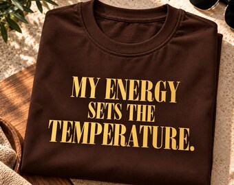 Può includere: Una t-shirt a maniche corte marrone scuro con il testo "MY ENERGY SETS THE TEMPERATURE." stampato in lettere maiuscole gialle e in grassetto al centro del petto. Il capo presenta un classico girocollo e un tessuto liscio e a tinta unita.