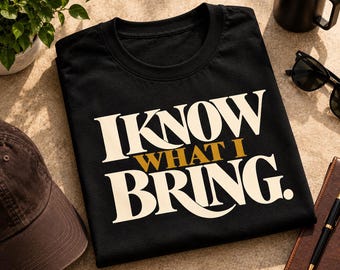 Può includere: Una t-shirt nera a maniche corte con il testo "I KNOW WHAT I BRING." in grandi lettere graziate color crema, con la parola "WHAT" stampata in un carattere più piccolo di colore oro, centrata tra le due righe di testo.