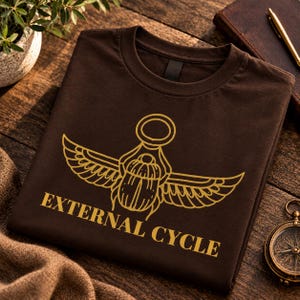 May include: A dark brown crew neck t-shirt featuring a gold-colored graphic of a winged scarab beetle centered above the text "EXTERNAL CYCLE" in a bold, capitalized font. The design is printed in a clean, minimalist line art style.
