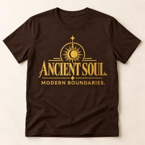 Op de afbeelding: Een donkerbruin t-shirt met korte mouwen en een gouden grafische print op de borst. Het ontwerp bevat een gestileerd zon- en maansikkel-embleem boven de tekst ANCIENT SOUL. en de tekst MODERN BOUNDARIES. in een strak serif-lettertype.