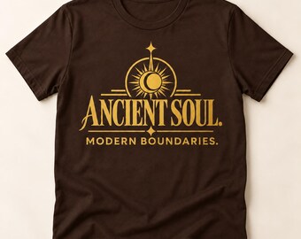 Può includere: Una t-shirt a maniche corte marrone scuro con una stampa grafica dorata sul petto. Il design include un emblema stilizzato di sole e luna crescente sopra il testo ANCIENT SOUL. e il testo MODERN BOUNDARIES. in un carattere serif pulito.