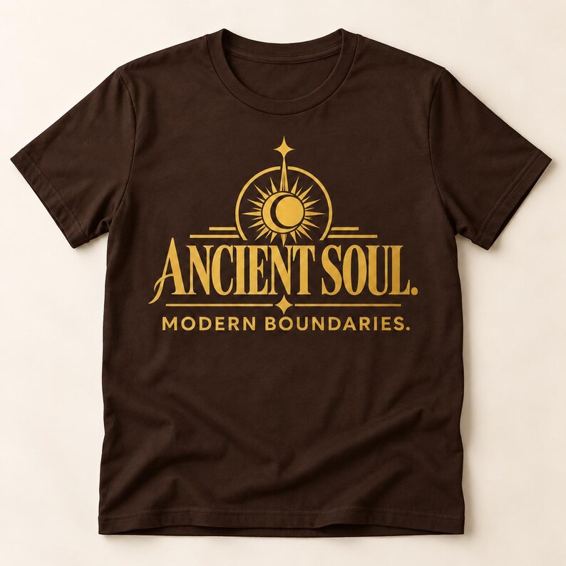 Op de afbeelding: Een donkerbruin t-shirt met korte mouwen en een gouden grafische print op de borst. Het ontwerp bevat een gestileerd zon- en maansikkel-embleem boven de tekst ANCIENT SOUL. en de tekst MODERN BOUNDARIES. in een strak serif-lettertype.
