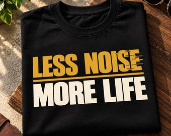 Può includere: Una t-shirt nera a girocollo con la scritta LESS NOISE in lettere dorate con un effetto di sfocatura da movimento sul lato destro, seguita da una linea orizzontale dorata e dalla scritta MORE LIFE in grandi lettere maiuscole bianche sul petto.