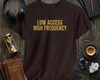 Può includere: Una t-shirt marrone scuro con il testo "LOW ACCESS HIGH FREQUENCY" in lettere dorate. Il capo è mostrato insieme a cuffie nere, una clessidra, un libro antico intitolato "The Philosophy of Poetry", un taccuino con penna e dei cristalli.