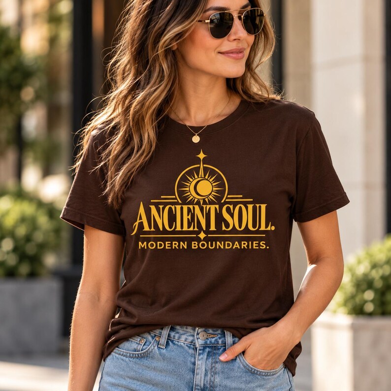 Op de afbeelding: Een donkerbruin T-shirt met korte mouwen en een gele grafische print op de borst. Het ontwerp bevat een zon- en maanicoon boven de tekst ANCIENT SOUL. en de tekst MODERN BOUNDARIES. in een strak, schreefloos lettertype eronder.