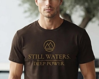 Può includere: Una t-shirt a maniche corte marrone scuro con una grafica dorata di due cime montuose all'interno di un cerchio. Sotto la grafica, il testo recita "STILL WATERS." seguito da una linea orizzontale e dal testo "DEEP POWER." in lettere dorate.
