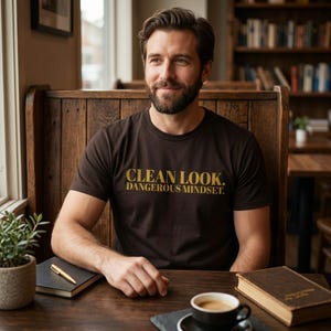 Op de afbeelding: Een donkerbruin T-shirt met korte mouwen en de tekst CLEAN LOOK. DANGEROUS MINDSET. in een goudkleurig lettertype op de borst. Het kledingstuk heeft een ronde hals en een standaard pasvorm, gemaakt van een effen gekleurde stof.