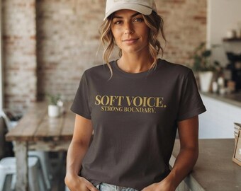 Può includere: Una t-shirt a maniche corte grigio scuro con la scritta dorata "SOFT VOICE. STRONG BOUNDARY." sul petto, abbinata a un berretto da baseball beige chiaro con un piccolo disegno ricamato sul pannello frontale sopra la visiera.