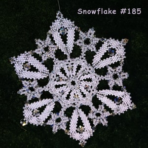 Puede incluir: Un adorno de copo de nieve blanco con cuentas y un centro de cristal. El copo de nieve está etiquetado como "Snowflake #185" y "Sandra D Halpenny".