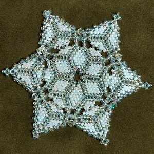 Puede incluir: Un adorno de copo de nieve de cuentas blancas y plateadas, sobre un fondo marrón. El adorno está diseñado por Sandra D Halpenny. Copo de nieve #194.