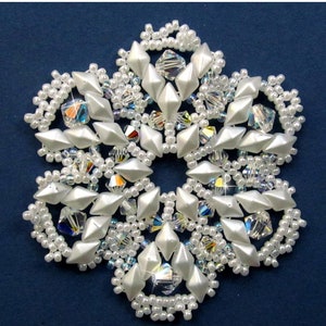 Puede incluir: Un patrón de adorno de copo de nieve blanco sobre un fondo azul. El patrón está hecho con cuentas y cristales. El texto "Snowflake #115 Ornament Pattern" está en la parte superior de la imagen y "Sandra D. Halpenny" en la parte inferior.