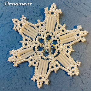 Puede incluir: Adorno de copo de nieve blanco con cuentas y un centro de cristal. El copo de nieve está etiquetado como "Snowflake #107 Ornament" y "Sandra D Halpenny".