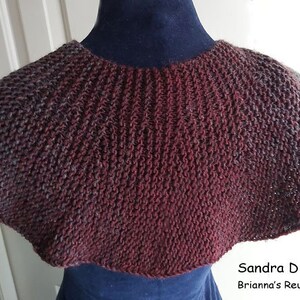 Peut inclure: Un capuchon en tricot bordeaux et gris avec un bord festonn&eacute;. Le capuchon est pr&eacute;sent&eacute; sur un mannequin. Le texte "Sandra D Halpenny Brianna's Reunion Capelet" est visible en bas de l'image.